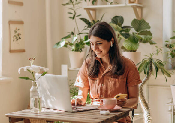 24-01 Positive young mixed race woman using a laptop and smartphone at home.Cozy home interior with indoor plants.Remote work, business,freelance,online shopping,e-learning,urban jungle concept.