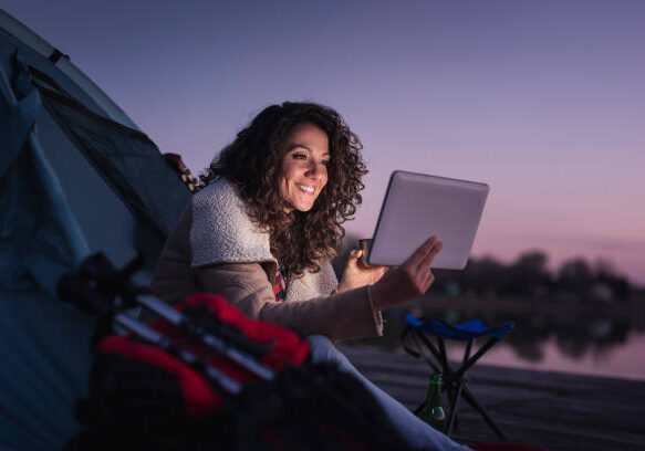 24-158 Young woman camping by the lake, sitting on the tent entrance, drinking hot coffee and reading the news on a tablet computer