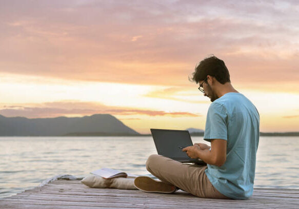 24-38 young digital nomad man sitting on wooden pier at sea working on internet remotely at sunset - Traveling with a computer - Online dream job concept