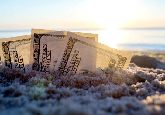 24-40 Three dollar bills are buried in sand on sandy beach near sea at sunset dawn in summer close-up. Dollar bills partially buried in sand